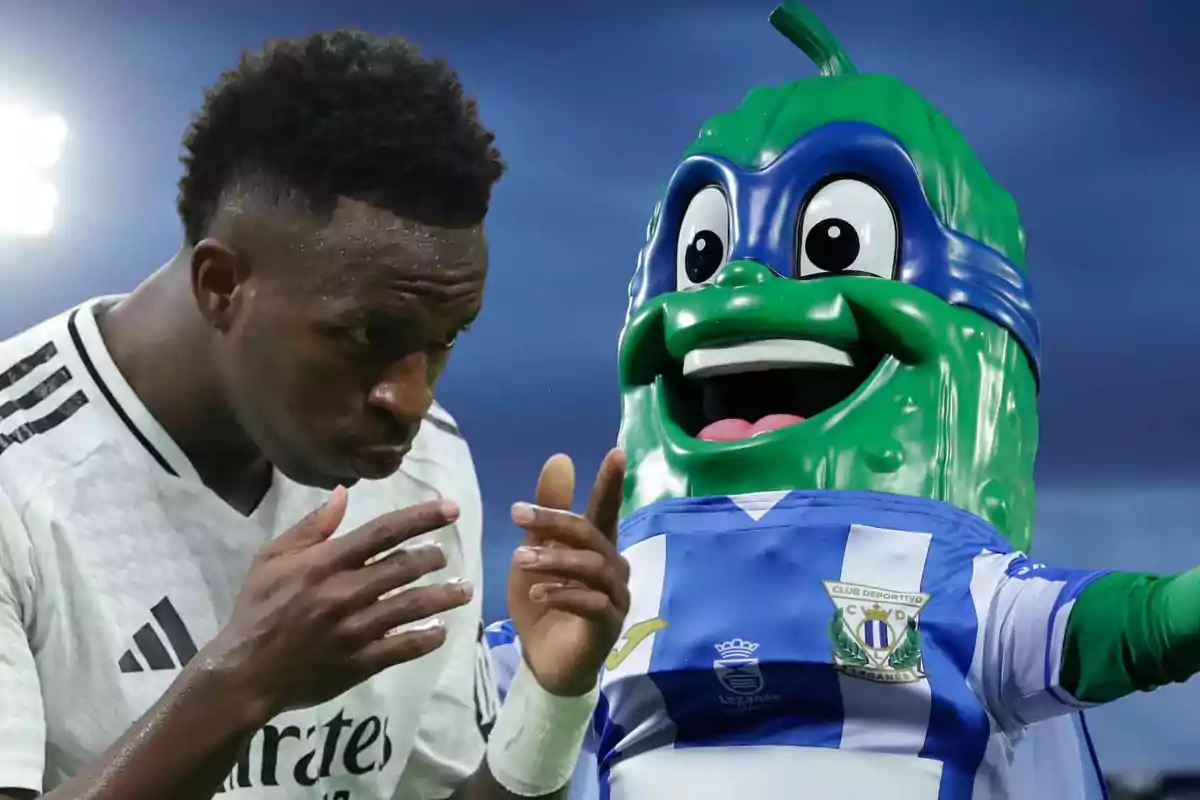 Un jugador de fútbol en uniforme blanco junto a una mascota verde con uniforme azul y blanco en un estadio.