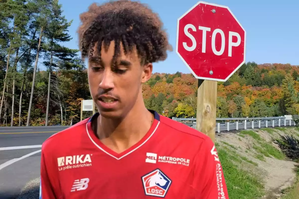 Un joven con una camiseta roja de un equipo de fútbol está de pie frente a una señal de stop en una carretera rodeada de árboles y vegetación otoñal.