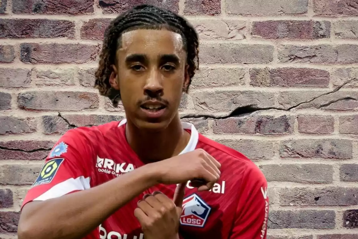 Un jugador de fútbol con una camiseta roja del equipo LOSC Lille posa frente a una pared de ladrillos agrietada.