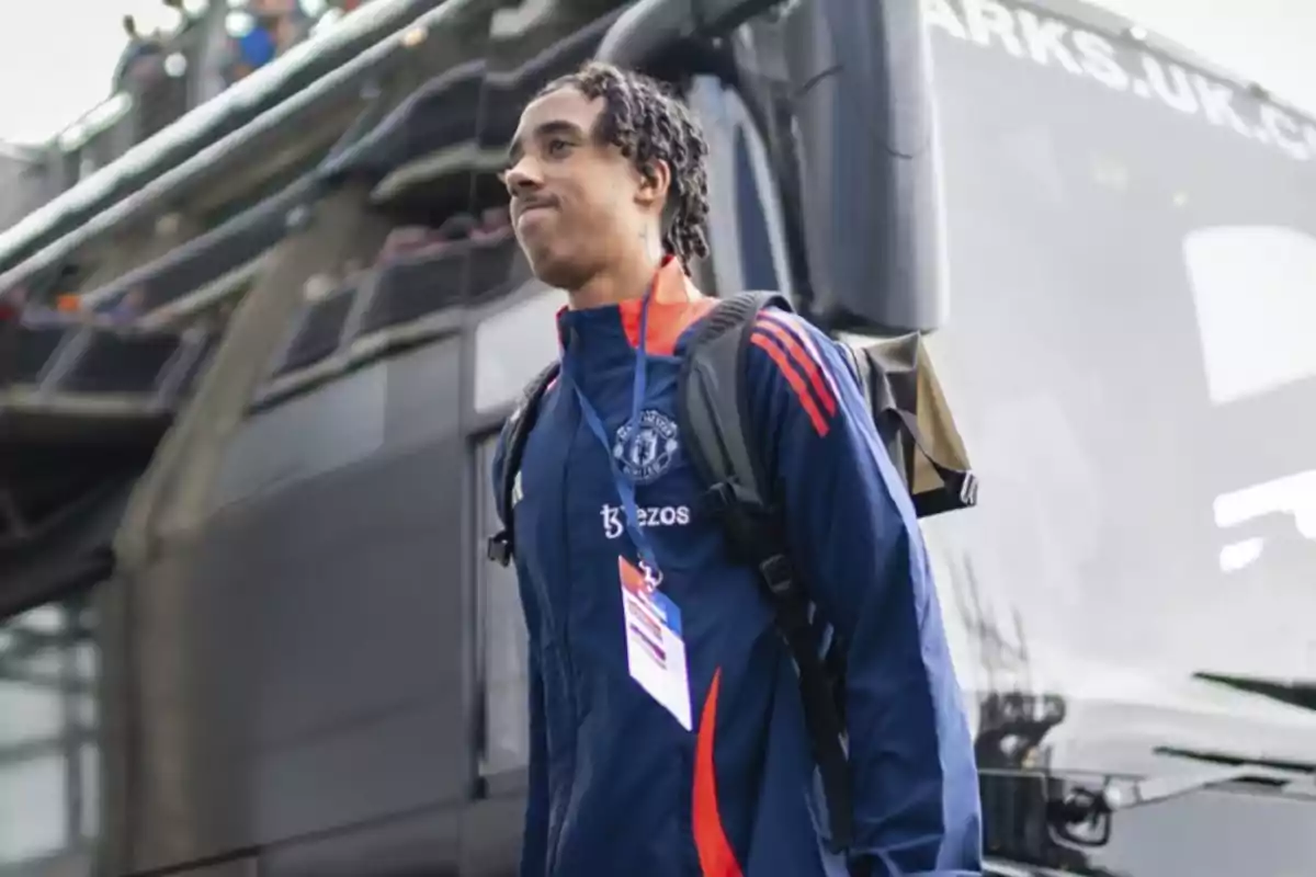 Un jugador de fútbol con uniforme azul y rojo del Manchester United, con una mochila, de pie frente a un autobús.