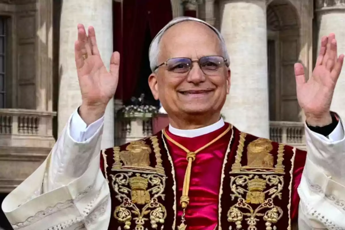 Un hombre mayor con gafas y vestimenta religiosa levanta las manos en señal de saludo frente a un edificio con columnas.