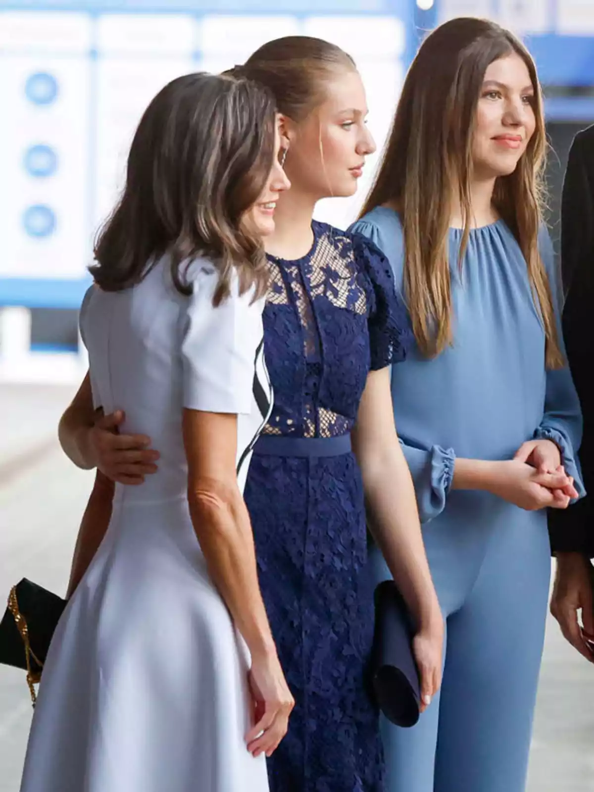 Tres mujeres posan juntas, una de ellas con un vestido blanco, otra con un vestido azul oscuro de encaje y la tercera con un mono azul claro.