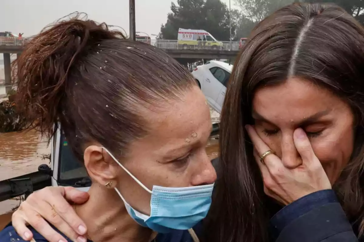 Dos mujeres se abrazan con expresiones de tristeza en un entorno afectado por inundaciones con un coche volcado y vehículos de emergencia al fondo.