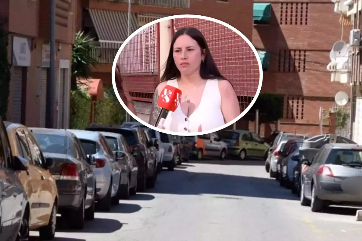 Una mujer sostiene un micrófono de un canal de televisión mientras está de pie en una calle con coches estacionados a ambos lados y edificios de ladrillo al fondo.
