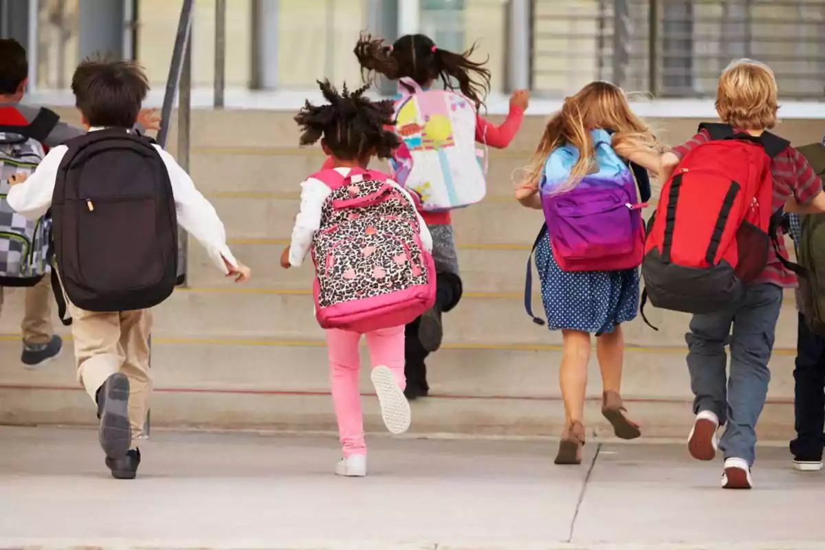 Niños corriendo hacia la entrada de la escuela con mochilas en la espalda.