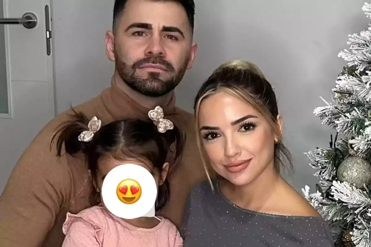 Una familia posando junto a un árbol de Navidad con la cara de la niña cubierta por un emoji de ojos de corazón. Una familia posando junto a un árbol de Navidad con la cara de la niña cubierta por un emoji de ojos de corazón.