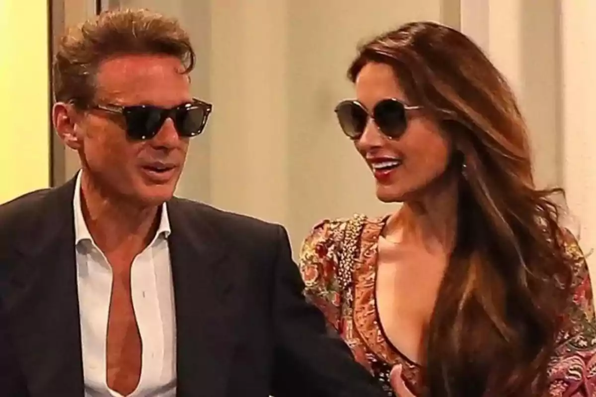 Una pareja sonriente con gafas de sol, el hombre lleva un traje oscuro y la mujer un vestido colorido. Una pareja sonriente con gafas de sol, el hombre lleva un traje oscuro y la mujer un vestido colorido.