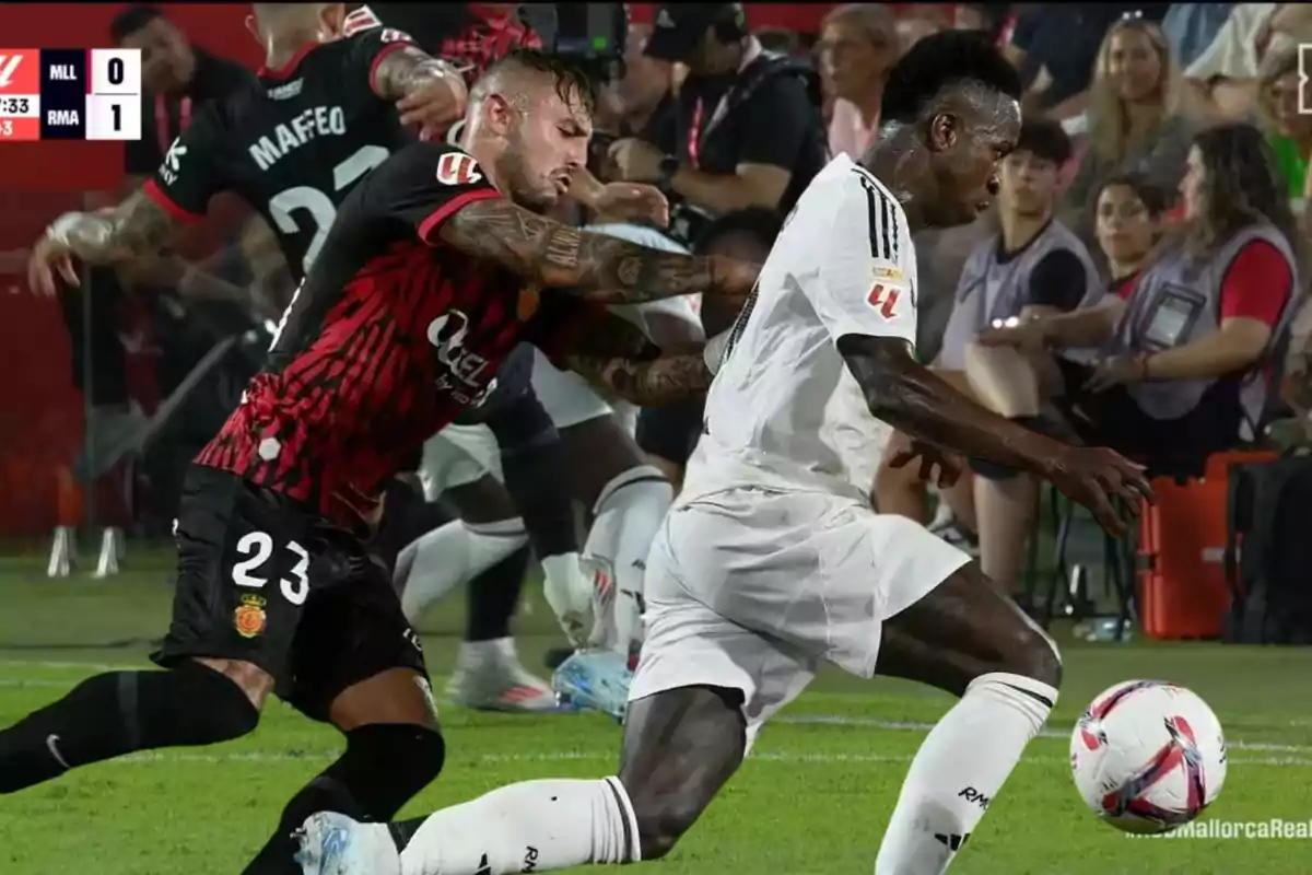 Un jugador del equipo con uniforme negro y rojo intenta detener a un jugador del equipo con uniforme blanco durante un partido de fútbol.