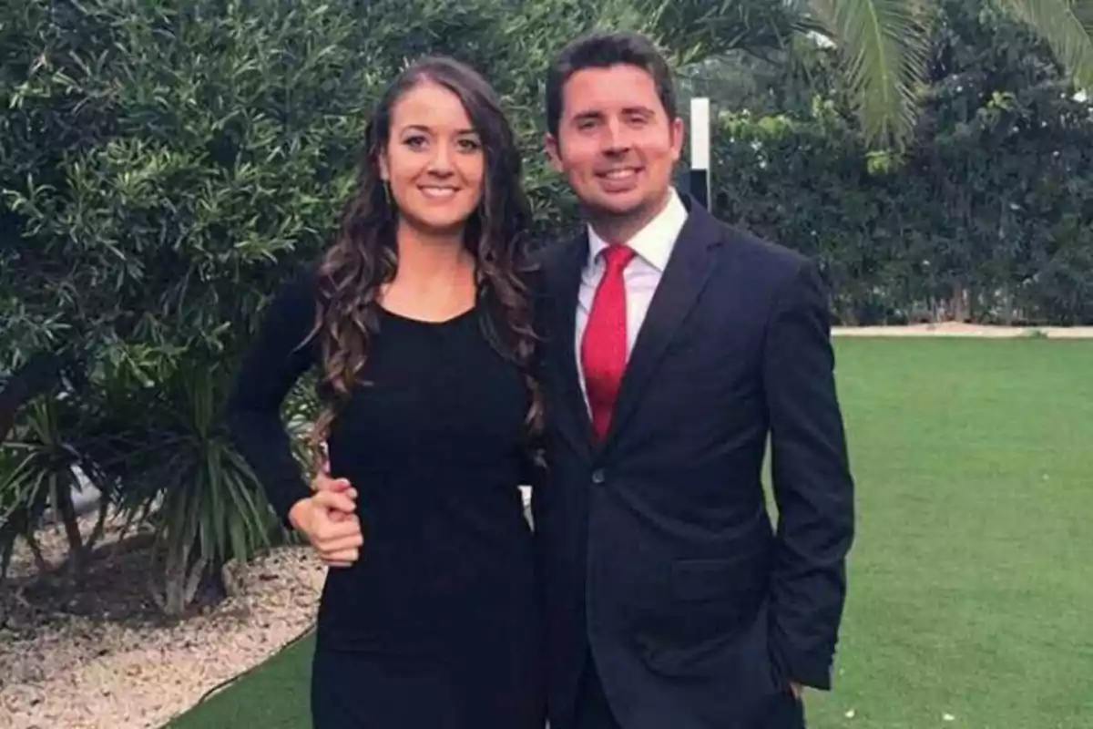 Pareja posando sonriente en un jardín, ella lleva un vestido negro y él un traje oscuro con corbata roja Pareja posando sonriente en un jardín, ella lleva un vestido negro y él un traje oscuro con corbata roja
