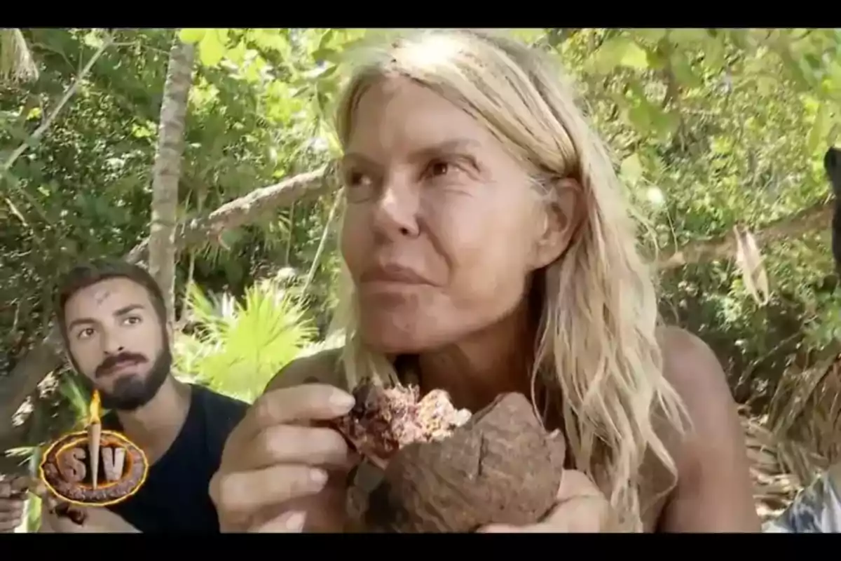 Una mujer rubia sostiene un coco mientras un hombre con barba la observa en un entorno selvático. Una mujer rubia sostiene un coco mientras un hombre con barba la observa en un entorno selvático.