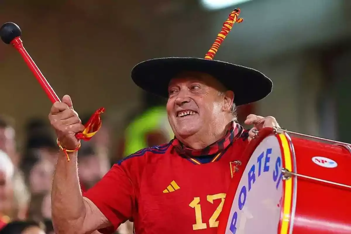 Un hombre sonriente con sombrero negro toca un tambor rojo en un evento deportivo. Un hombre sonriente con sombrero negro toca un tambor rojo en un evento deportivo.