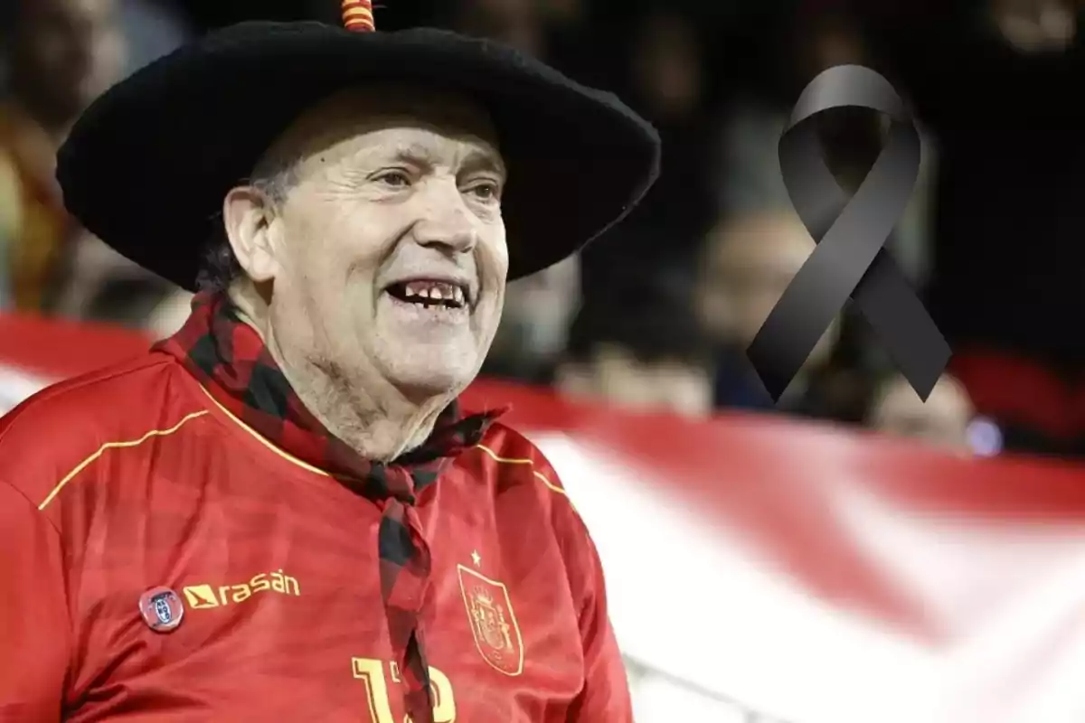 Hombre mayor con camiseta roja y sombrero negro sonríe en un evento deportivo con un lazo negro en señal de luto.