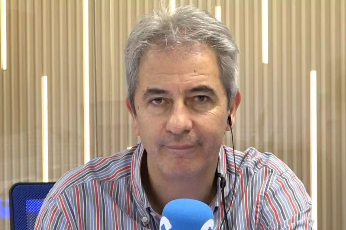 Hombre de cabello canoso con camisa a rayas y auriculares frente a un micrófono azul en un estudio.