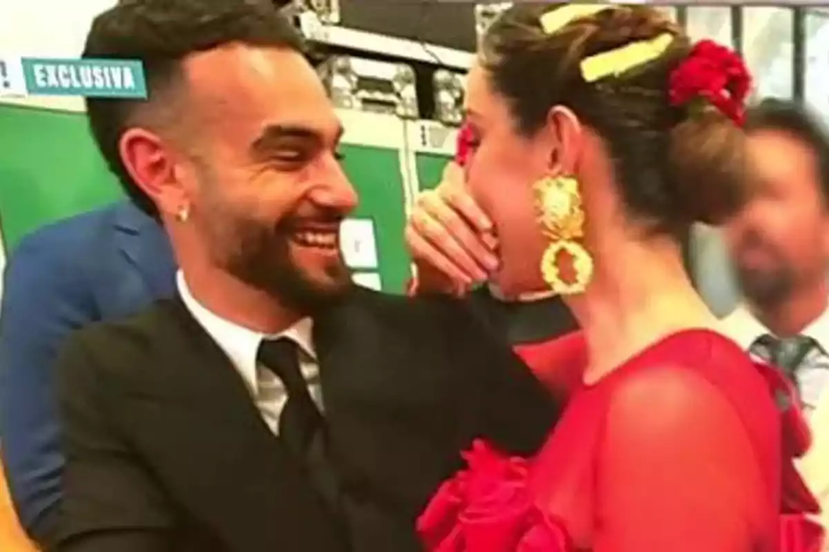 Una pareja sonriente comparte un momento de complicidad en un evento, ella lleva un vestido rojo y accesorios dorados mientras él viste un traje oscuro. Una pareja sonriente comparte un momento de complicidad en un evento, ella lleva un vestido rojo y accesorios dorados mientras él viste un traje oscuro.