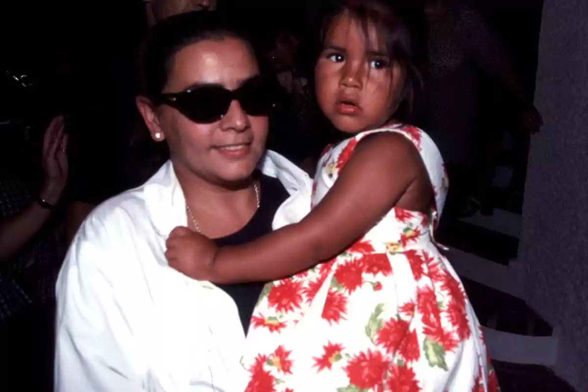 Una mujer con gafas de sol sostiene a una niña que lleva un vestido con estampado floral.