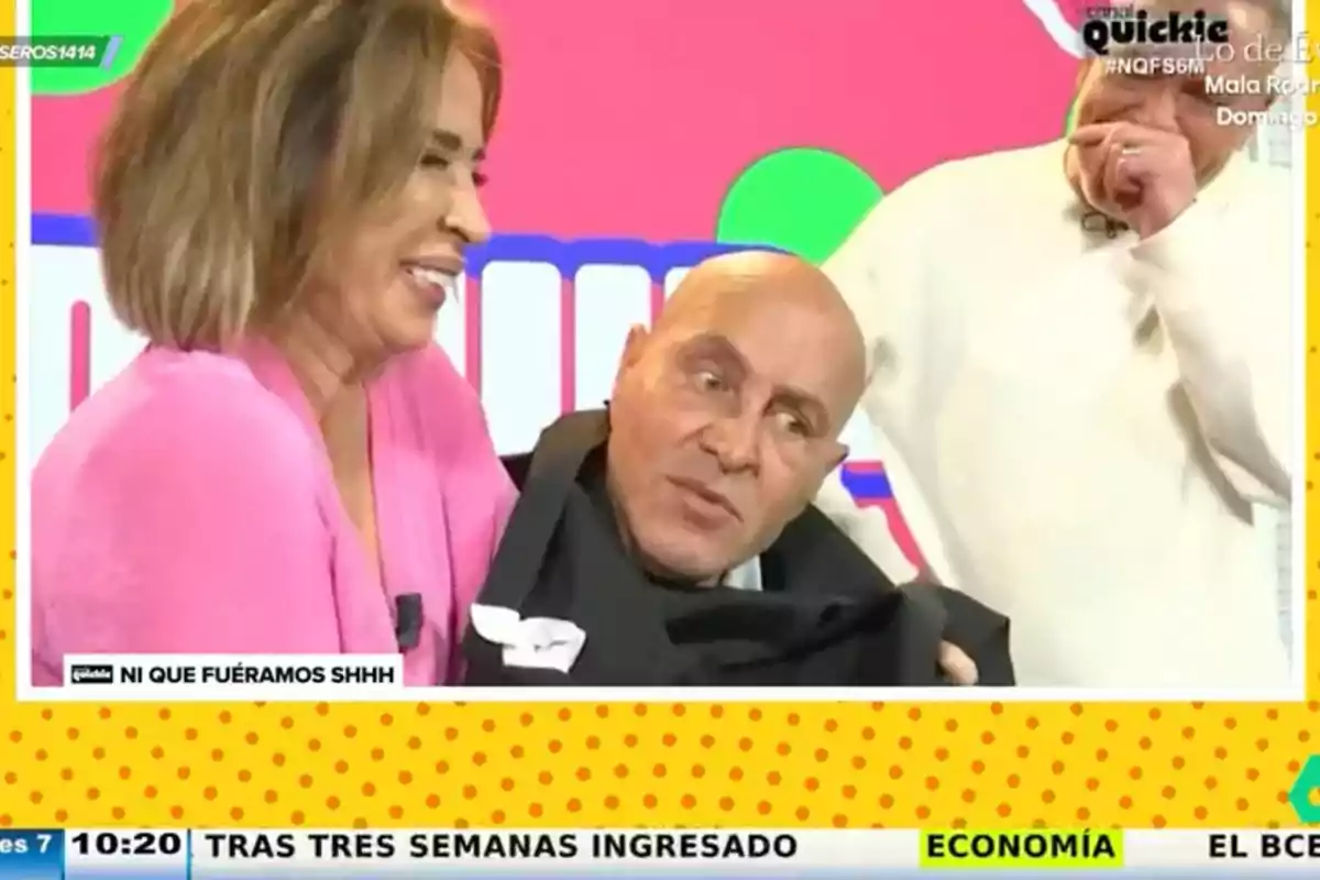 Una mujer sonríe mientras sostiene a un hombre que parece estar envuelto en una tela negra, con un fondo colorido y un marco amarillo con puntos. Una mujer sonríe mientras sostiene a un hombre que parece estar envuelto en una tela negra, con un fondo colorido y un marco amarillo con puntos.