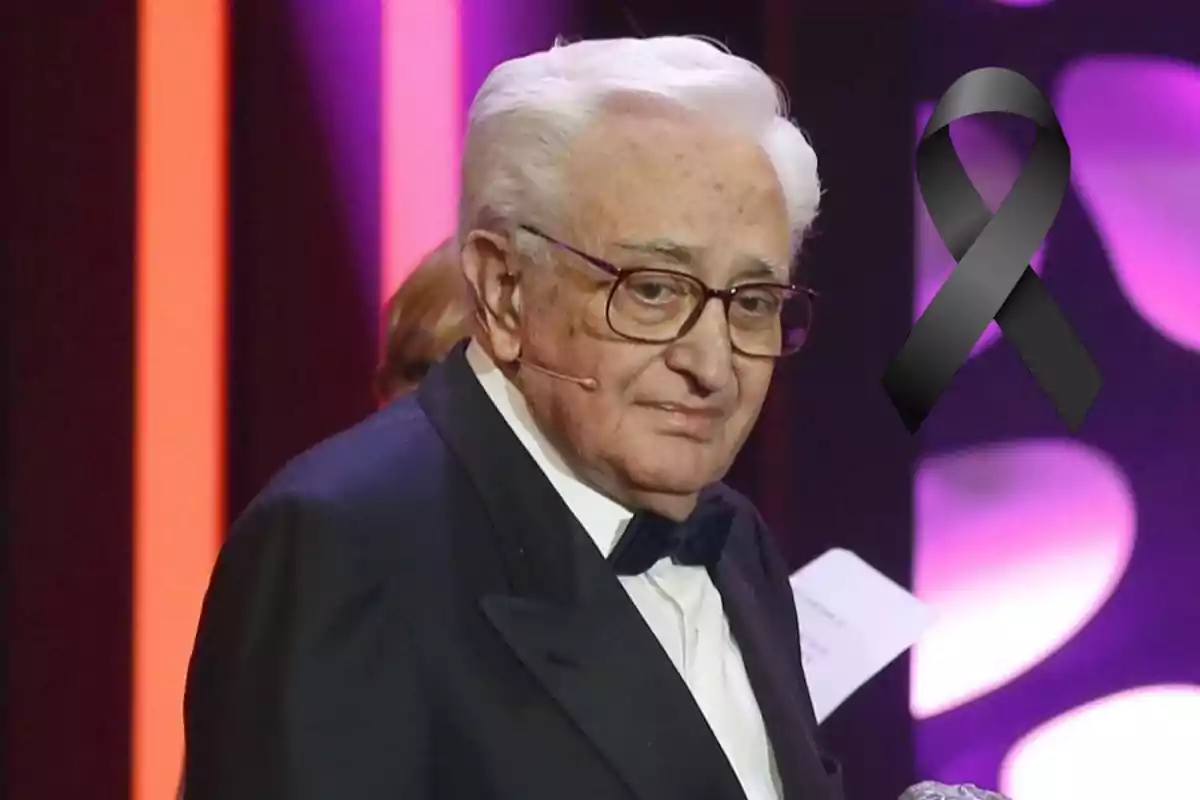 Un hombre mayor con gafas y esmoquin en un escenario con un lazo negro superpuesto en la imagen.