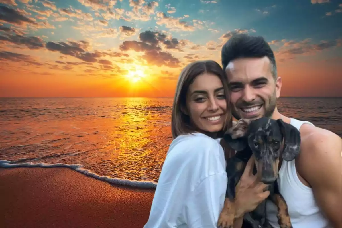 Una pareja sonriente con un perro en brazos posando frente a un atardecer en la playa.