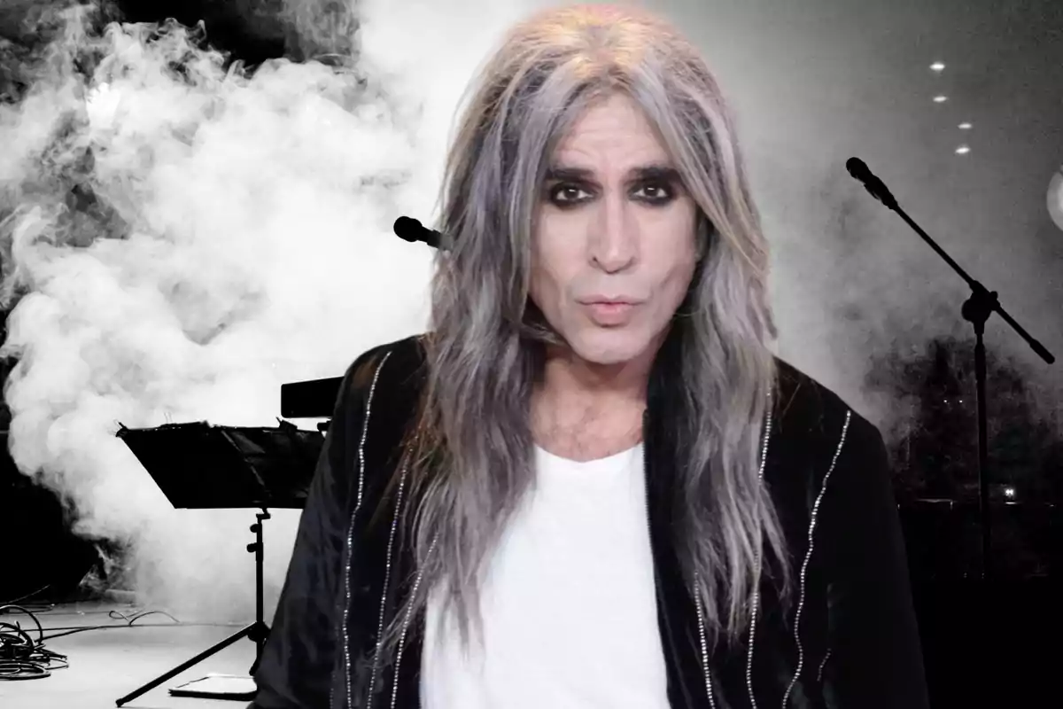 Un hombre de cabello largo y canoso con maquillaje oscuro en los ojos, vistiendo una chaqueta negra y camiseta blanca, posando frente a un fondo con humo y micrófonos.