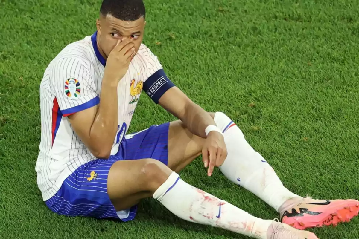 Jugador de fútbol sentado en el césped con uniforme blanco y azul, tocándose la cara y con una venda en la muñeca.