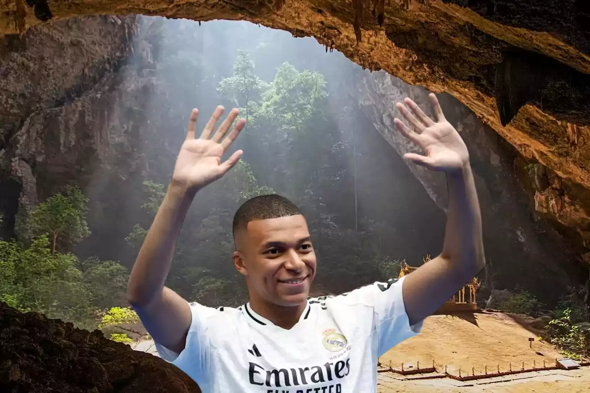 Un hombre con una camiseta blanca levantando las manos dentro de una cueva con vegetación y luz natural.