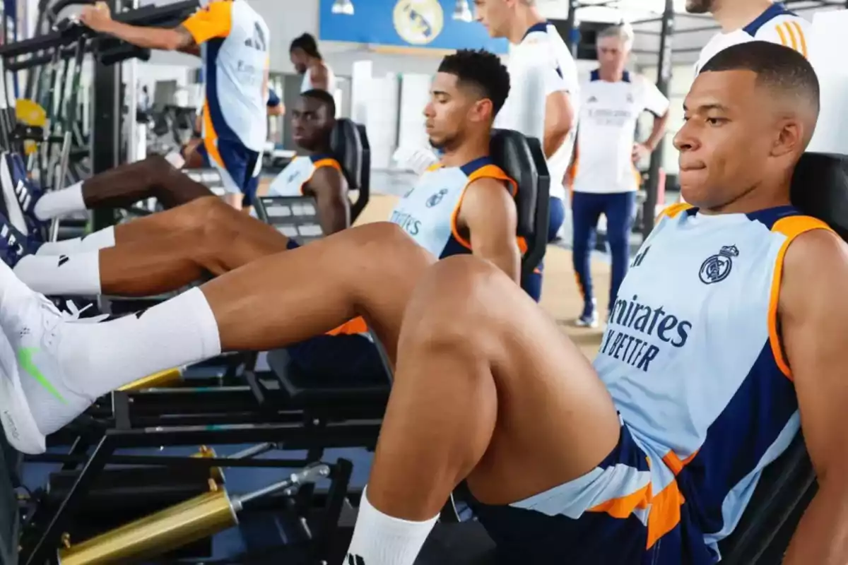 Jugadores de fútbol entrenando en el gimnasio, realizando ejercicios de piernas con máquinas de pesas, mientras visten uniformes de entrenamiento del Real Madrid.