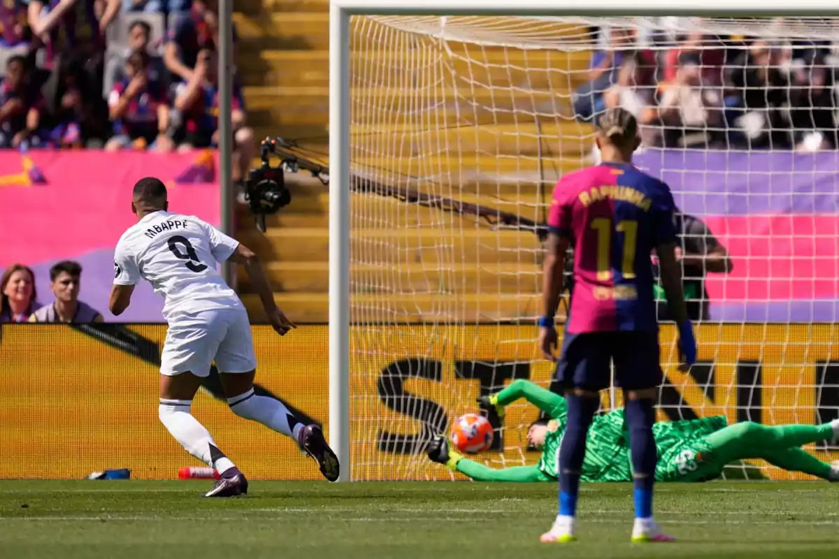 Un jugador de fútbol con el número 9 en su camiseta está a punto de patear un balón hacia la portería mientras el portero se lanza para intentar detenerlo y otro jugador observa la acción desde cerca. Un jugador de fútbol con el número 9 en su camiseta está a punto de patear un balón hacia la portería mientras el portero se lanza para intentar detenerlo y otro jugador observa la acción desde cerca.