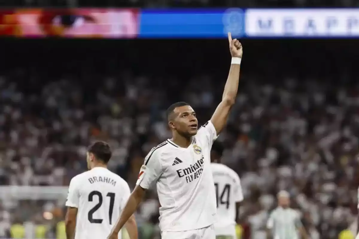 Un jugador de fútbol del Real Madrid levantando el brazo en señal de celebración durante un partido, con otros jugadores y el público de fondo.