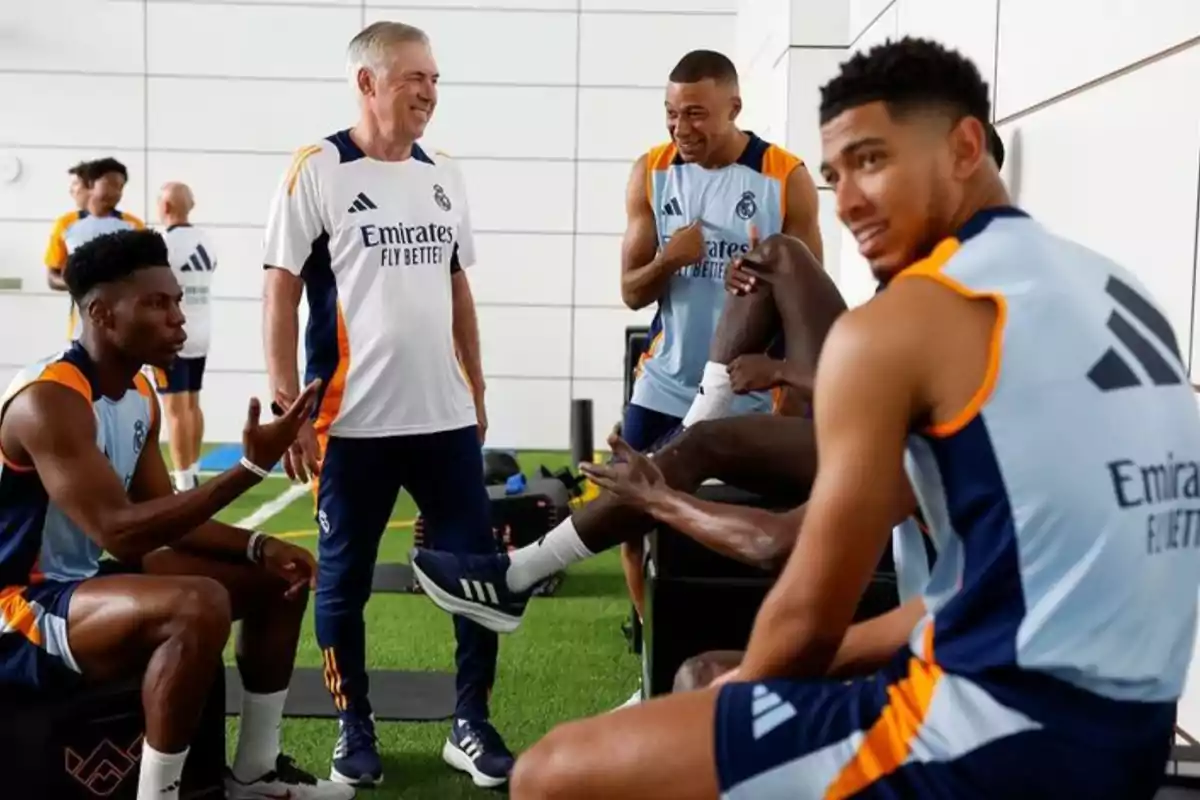 Un grupo de jugadores de fútbol y su entrenador están conversando y riendo en una sala de entrenamiento, todos llevan uniformes del Real Madrid.