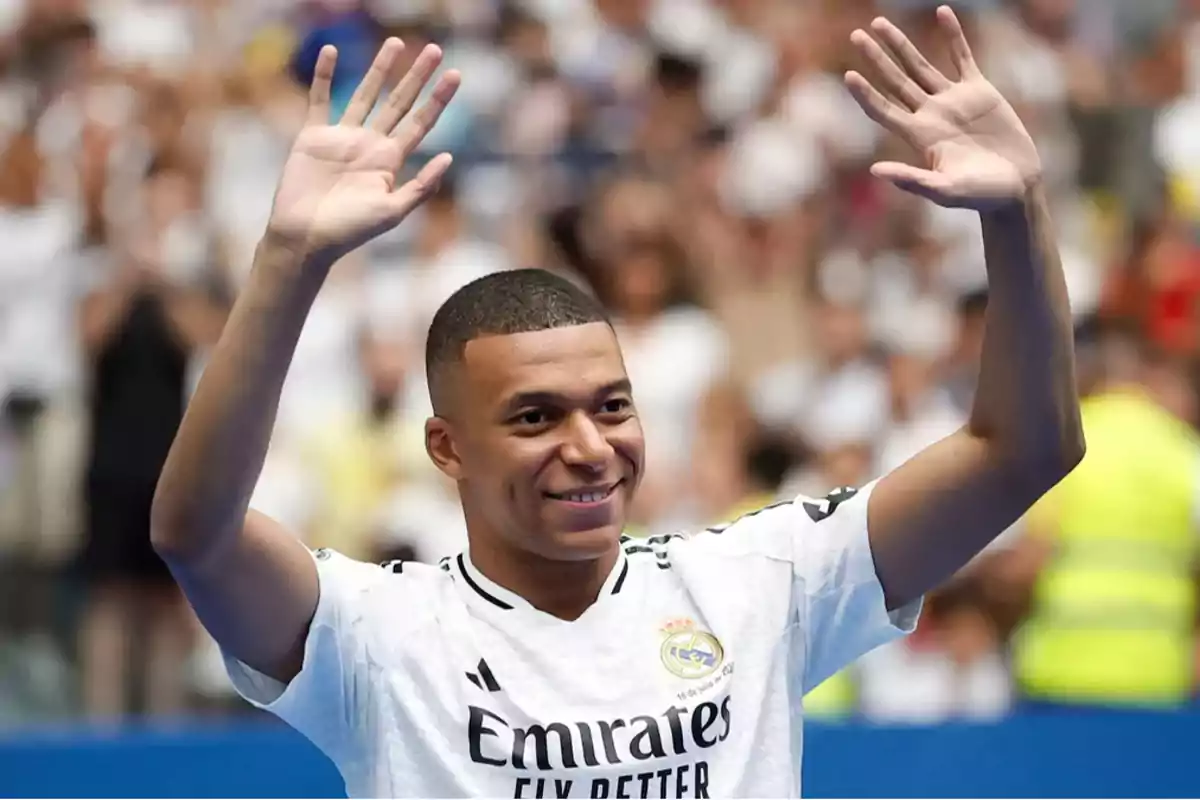 Mbappé con la camiseta del Real Madrid levantando las manos y sonriendo en un estadio lleno de aficionados.