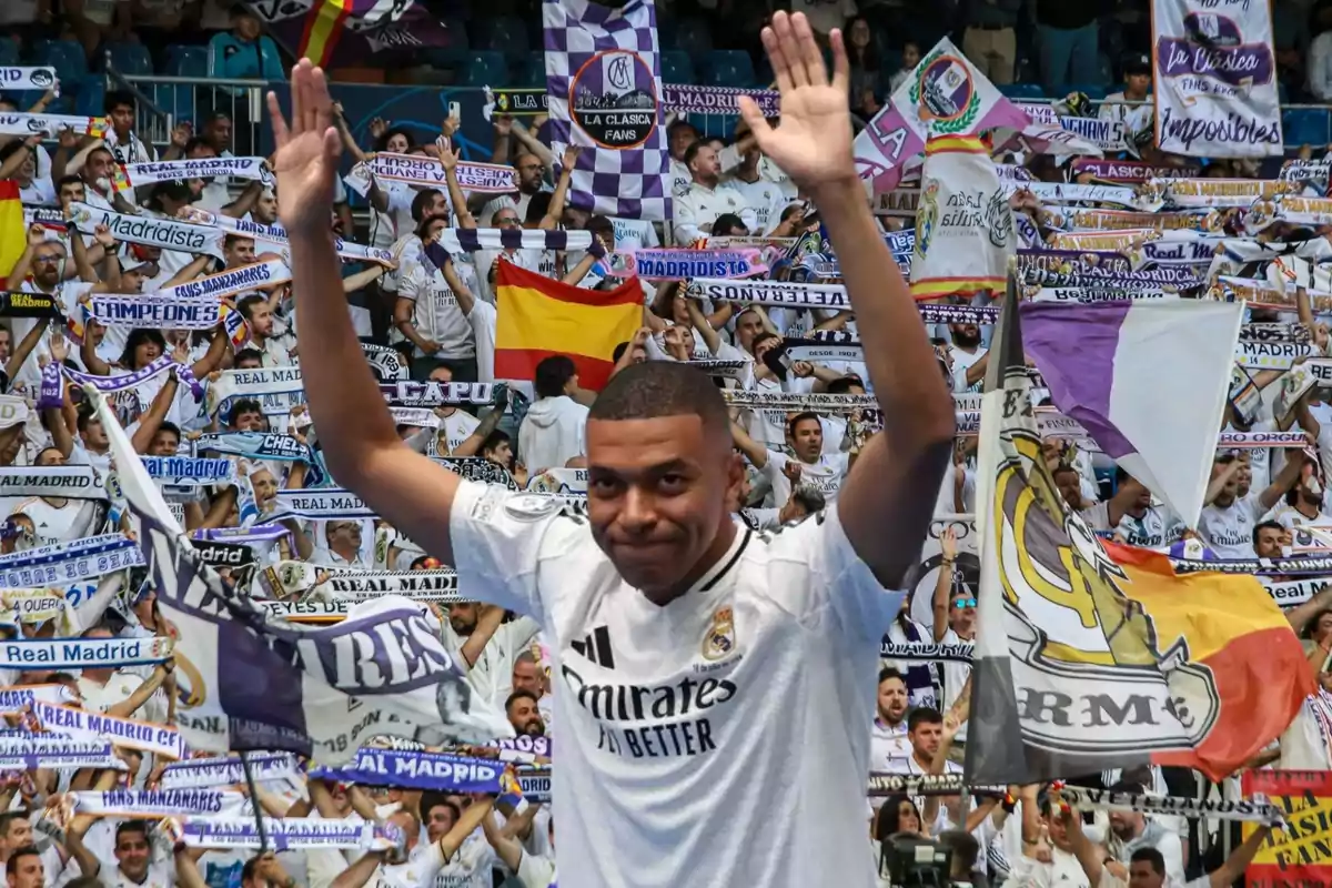 Un jugador de fútbol con la camiseta del Real Madrid saluda a una multitud de aficionados que sostienen banderas y bufandas del equipo.