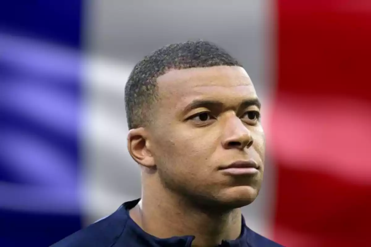 Mbappé con expresión seria frente a una bandera de Francia desenfocada.