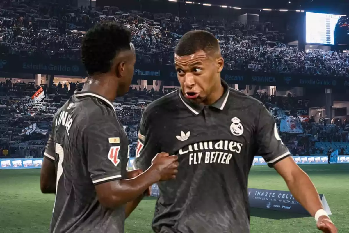 Dos jugadores de fútbol con uniformes oscuros conversan en un estadio lleno de espectadores.