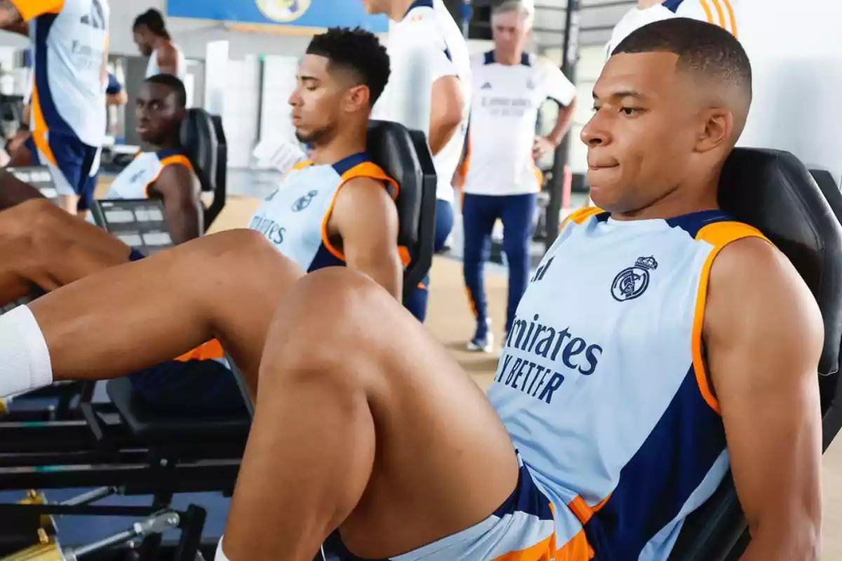 Jugadores de fútbol entrenando en el gimnasio, usando uniformes de entrenamiento del Real Madrid y realizando ejercicios de piernas en una máquina de pesas.