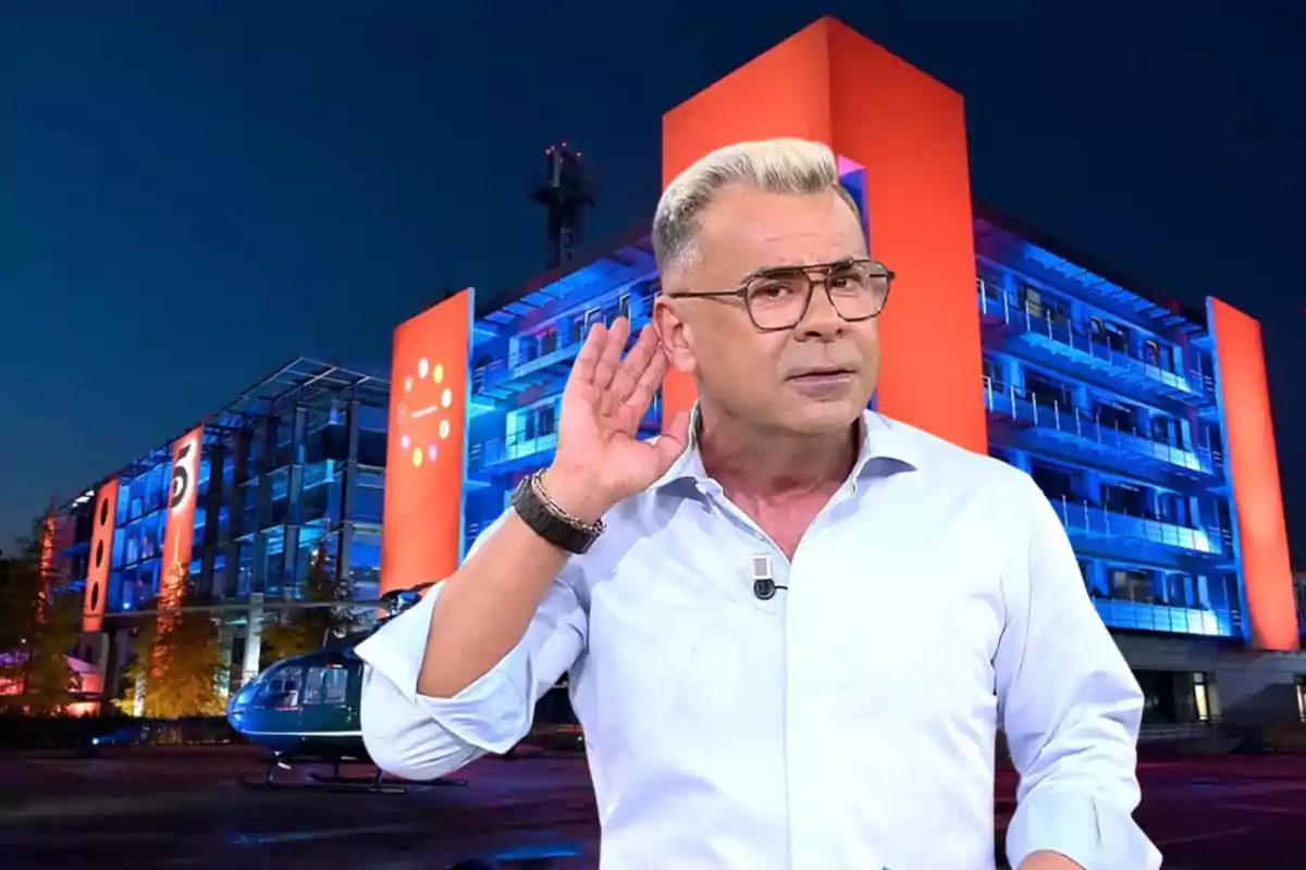 Hombre de cabello rubio y gafas con camisa clara haciendo un gesto de escuchar con la mano en la oreja frente a un edificio iluminado en azul y naranja