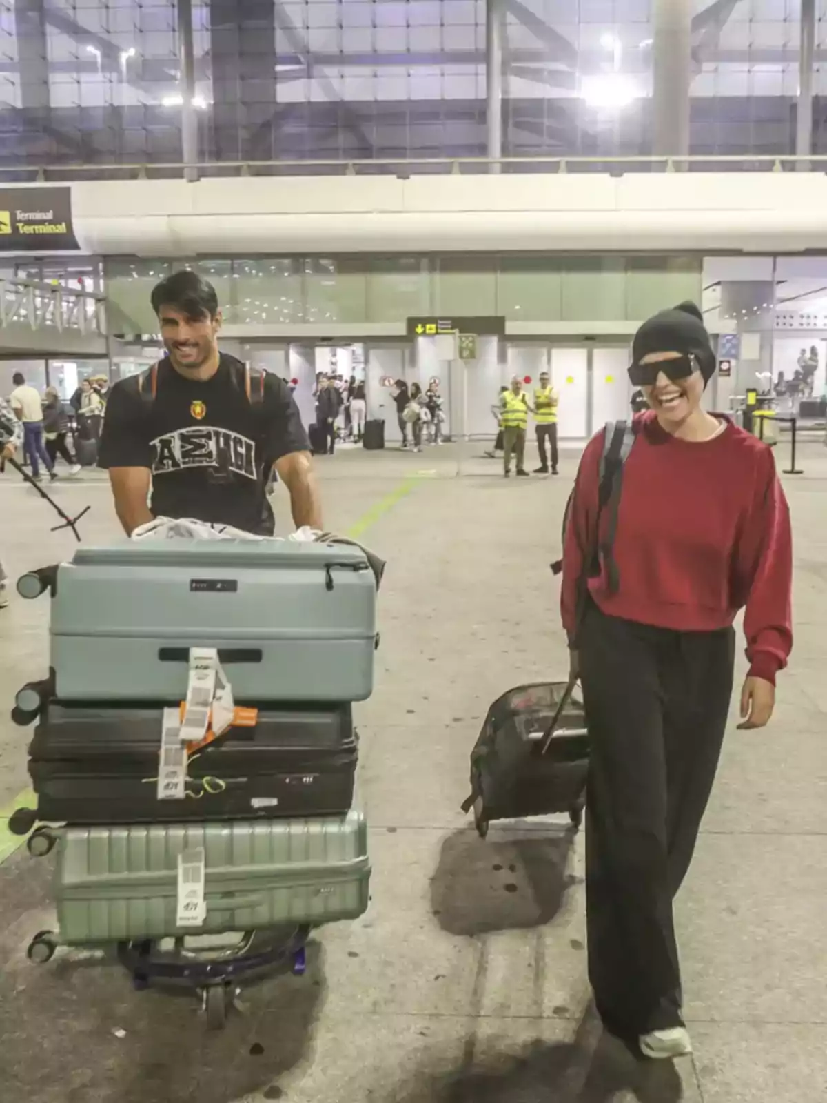 Una pareja sonriente camina por el aeropuerto, él empuja un carrito lleno de maletas y ella lleva una pequeña maleta de mano. Una pareja sonriente camina por el aeropuerto, él empuja un carrito lleno de maletas y ella lleva una pequeña maleta de mano.