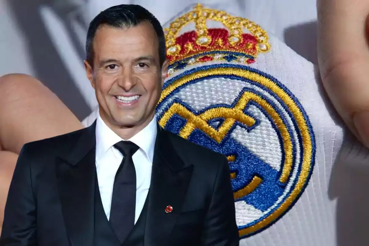 Un hombre con traje negro y corbata negra sonriendo frente a un escudo del Real Madrid.