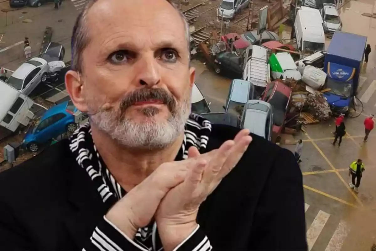 Miguel bosé en primer plano vestido de negro y de fondo varios coches destrozados y apilados