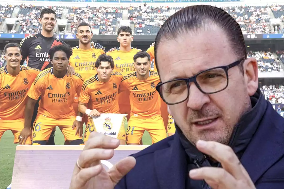 Un grupo de jugadores de fútbol con uniformes naranjas posan para una foto en el campo, mientras un hombre con gafas y chaqueta oscura aparece en primer plano gesticulando con las manos.