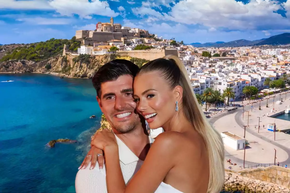 Una pareja sonriente se abraza frente a un paisaje costero con una ciudad y un castillo en el fondo.