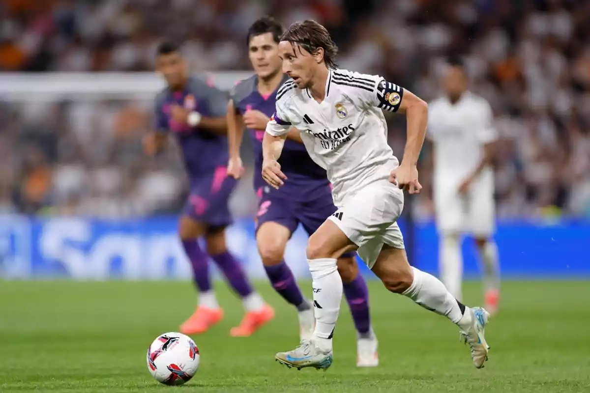 Jugador de fútbol del Real Madrid corriendo con el balón durante un partido, con jugadores del equipo contrario en el fondo.