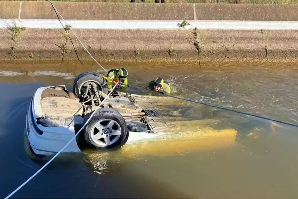 Un coche volcado en un canal de agua con dos personas en trajes de buceo trabajando para recuperarlo. Un coche volcado en un canal de agua con dos personas en trajes de buceo trabajando para recuperarlo.