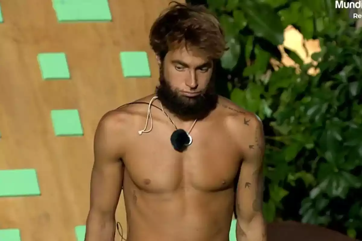 Hombre joven sin camiseta con barba espesa y expresión seria, lleva un micrófono colgado al cuello y está al aire libre frente a un fondo de plantas y una estructura de madera con cuadros verdes Hombre joven sin camiseta con barba espesa y expresión seria, lleva un micrófono colgado al cuello y está al aire libre frente a un fondo de plantas y una estructura de madera con cuadros verdes