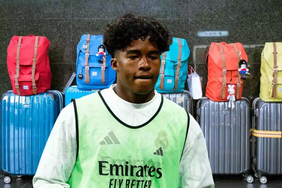 Un joven con un chaleco de entrenamiento del Real Madrid está frente a varias maletas y mochilas de colores brillantes.