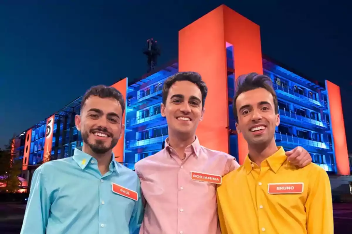 Tres personas sonrientes posan frente a un edificio iluminado con luces azules y naranjas.