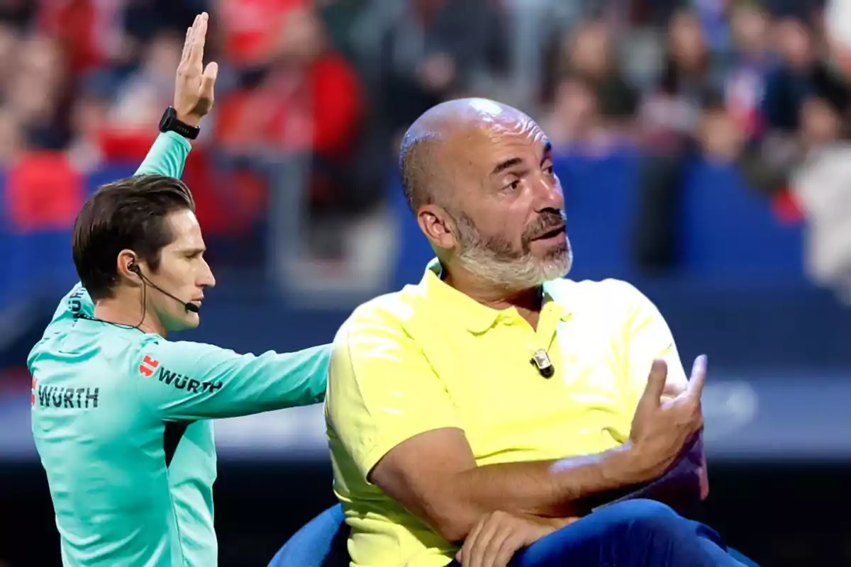 Un árbitro de fútbol levanta la mano mientras un hombre con camisa amarilla está sentado y gesticulando.