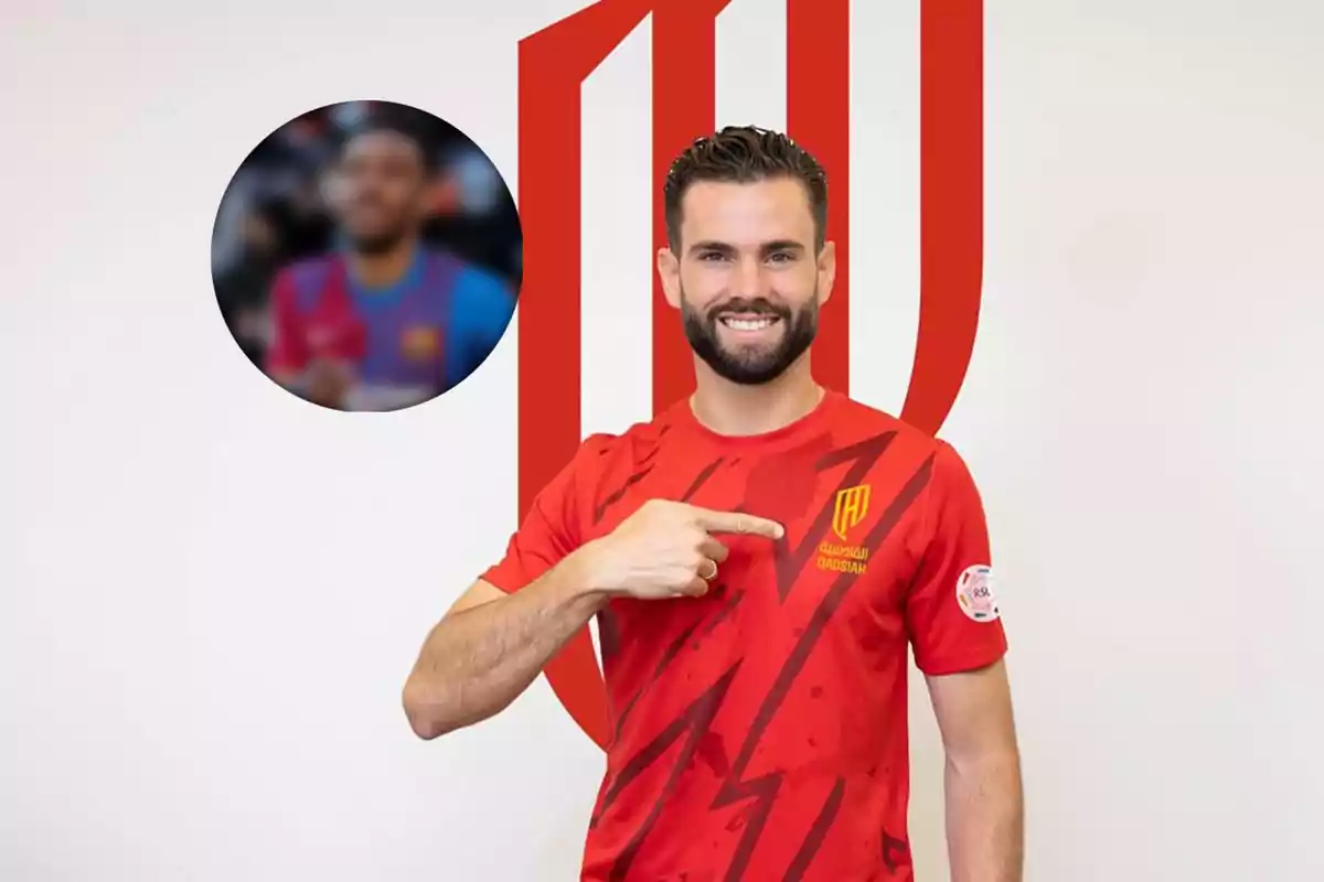 Un hombre sonriente con barba y cabello corto, vestido con una camiseta roja con el logo de Al-Qadsiah, señala el escudo del equipo mientras posa frente a un fondo blanco con un gran logotipo rojo.