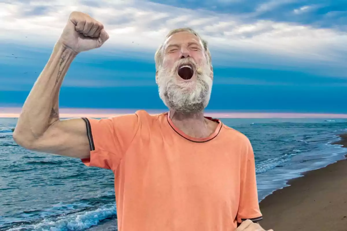 Hombre mayor con barba blanca y camiseta naranja levantando el puño y gritando en una playa con el mar y el cielo azul de fondo.