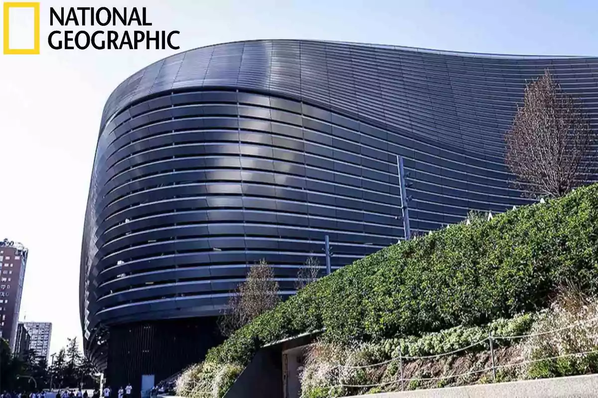 Edificio moderno con fachada curva y líneas horizontales junto a un jardín verde bajo un cielo despejado.