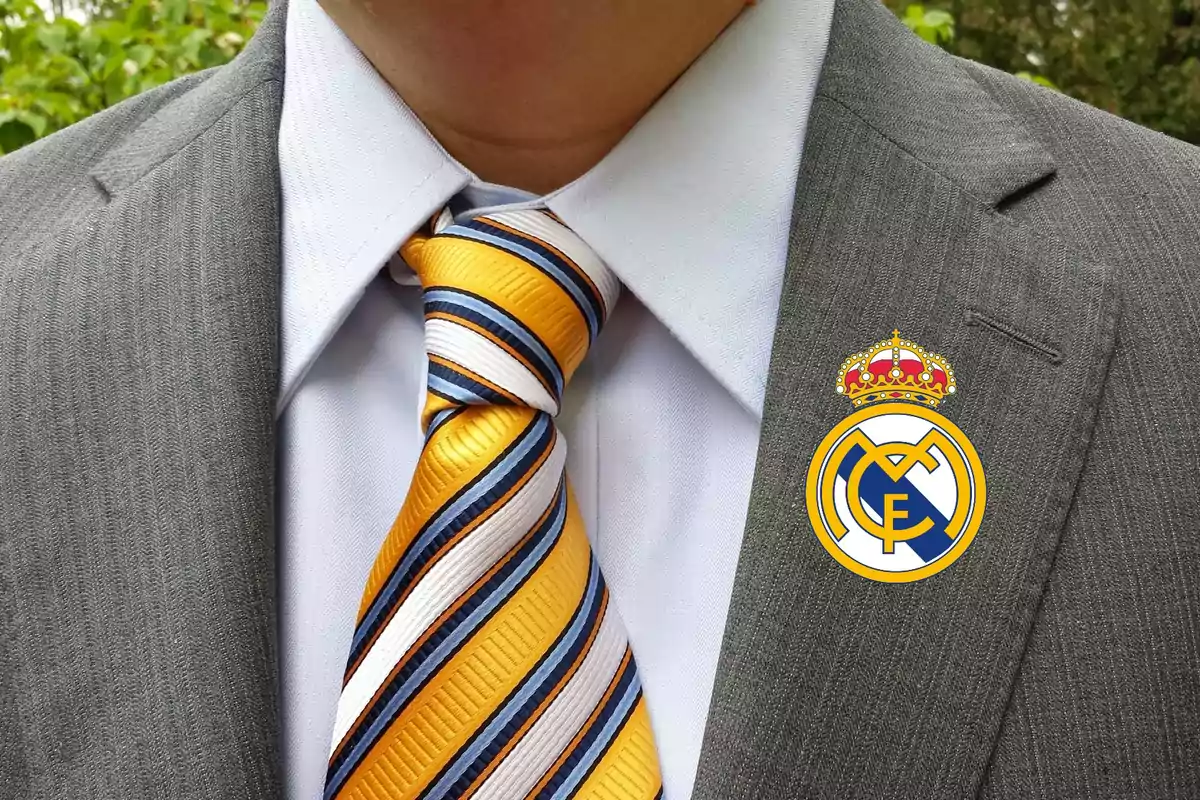 Un hombre con traje gris, camisa blanca y corbata a rayas amarillas, blancas y azules, con el escudo del Real Madrid en la solapa del saco.